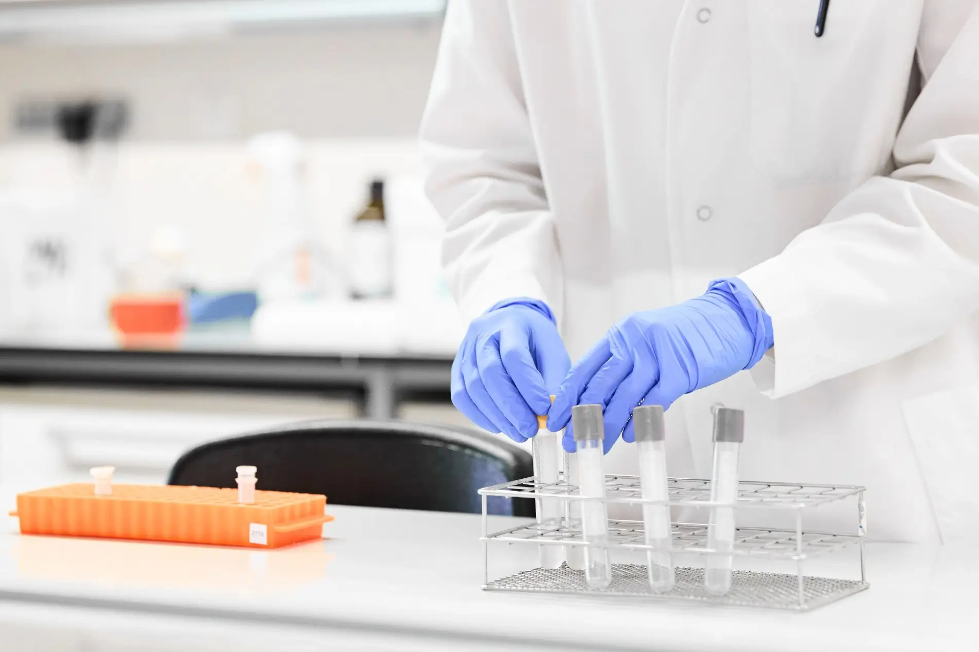 A person in a white lab coat and blue gloves places a test tube into a rack