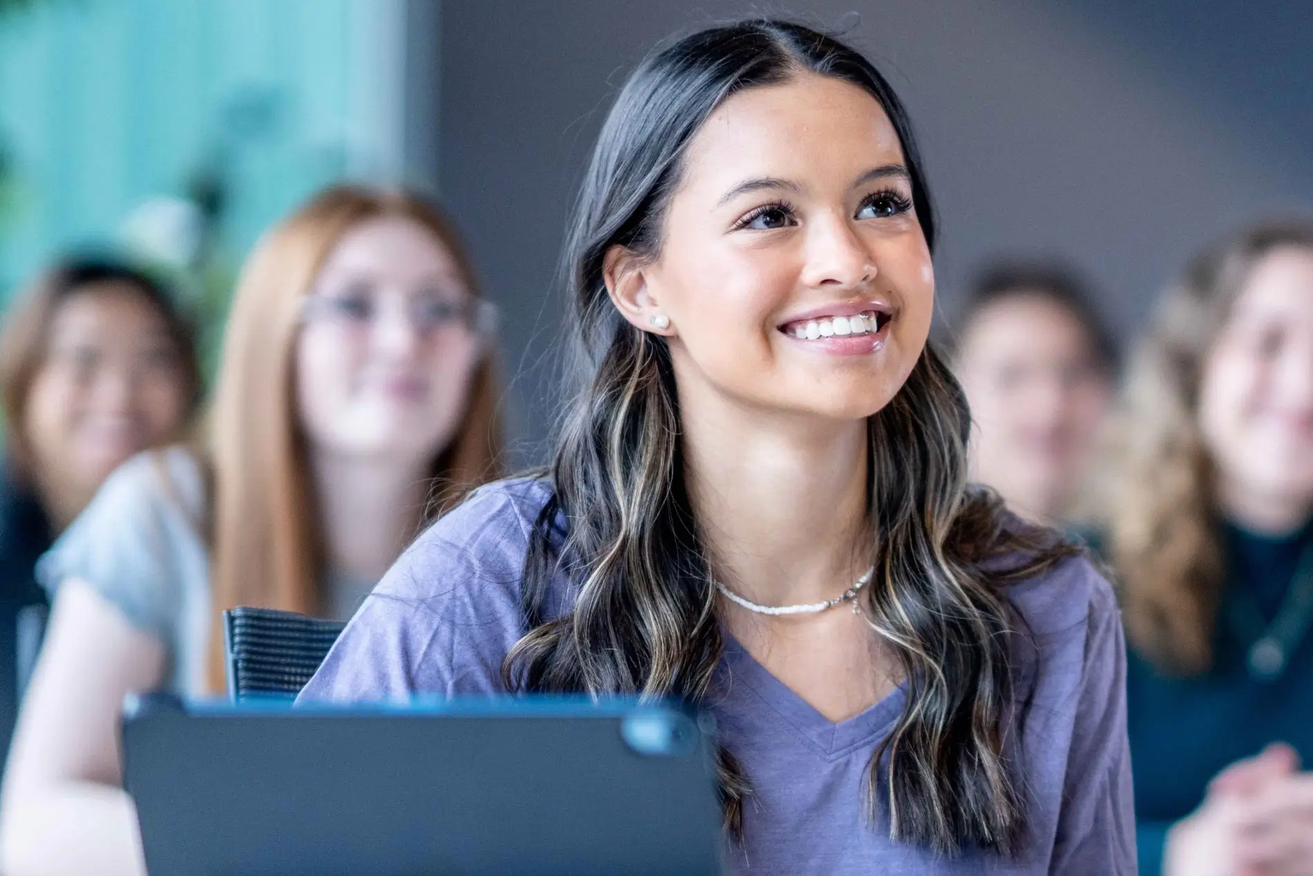 A young female student with long brown hair smiles brightly, looking up white sitting at a desk. Other students are blurred in the background.