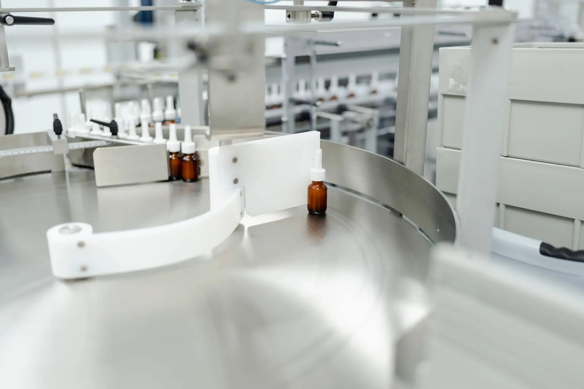 A small brown bottle moves along a clean, metallic conveyor belt in a pharmaceutical manufacturing facility.