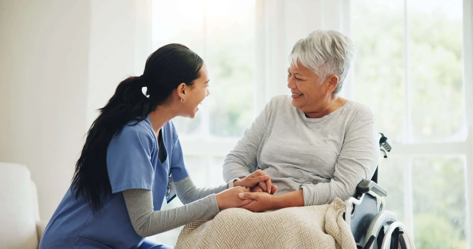 A healthcare professional kneels beside an elderly woman in a wheelchair, holding her hands.