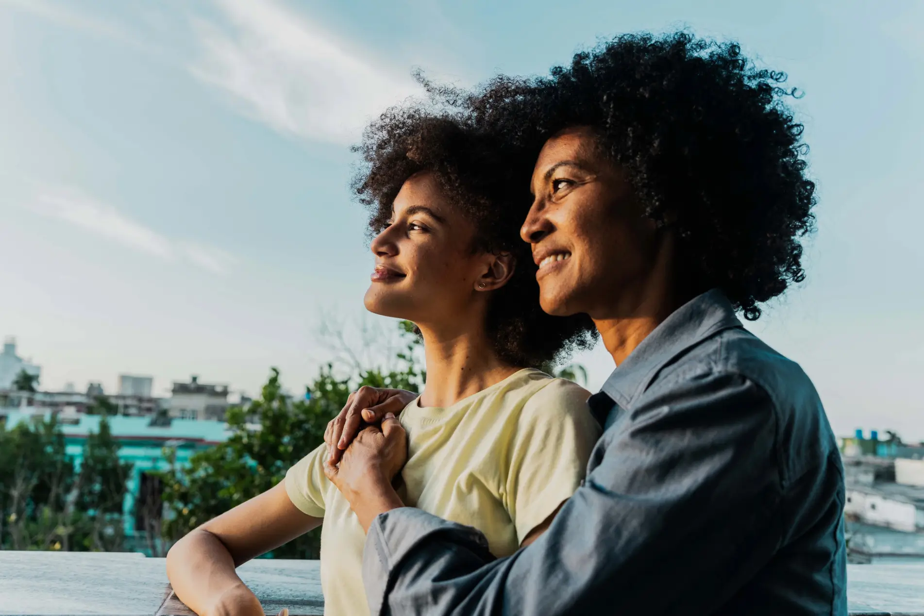 A younger woman and an older woman with curly hair stand outdoors, smiling and looking into the distance.
