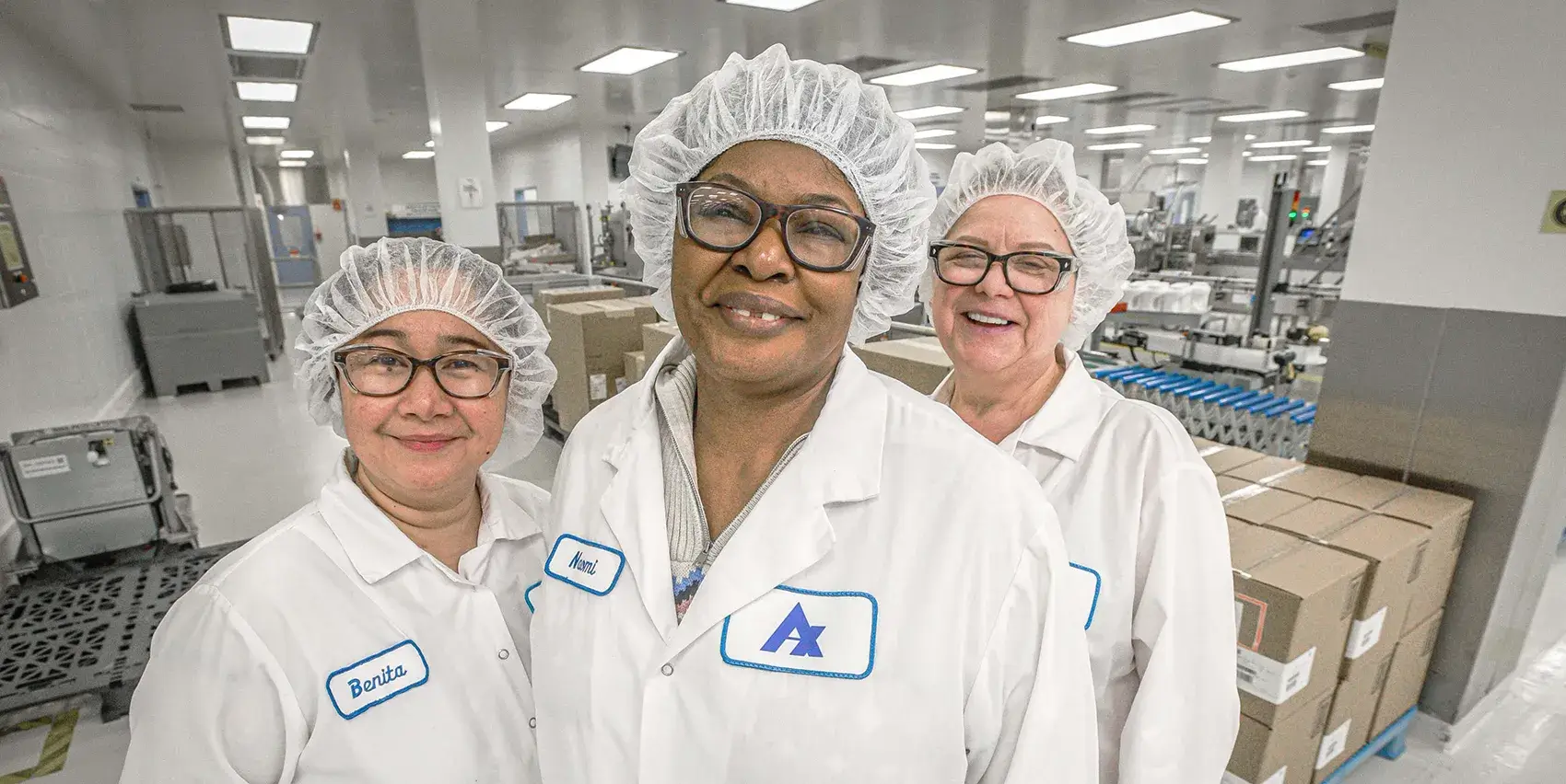Three women in white lab coats and hairnets smile in a clean room facility.